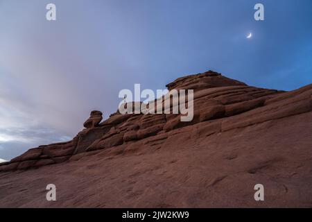 D'en-dessous de touristes se tenant sur le sommet de la formation de Rock équilibré contre le ciel du soir avec la lune dans le parc national d'Arches dans Utah, États-Unis Banque D'Images