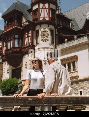Jeune couple aimant positif en vêtements décontractés et lunettes de soleil souriant et regardant loin tout en se tenant près du vieux château de Peles le jour ensoleillé en Roumanie Banque D'Images