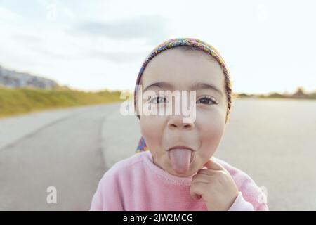 Adorable préchooleur hispanique en sweat-shirt et foulard rose regardant l'appareil photo et montrant la langue tout en se tenant sur la route asphaltée par temps nuageux Banque D'Images