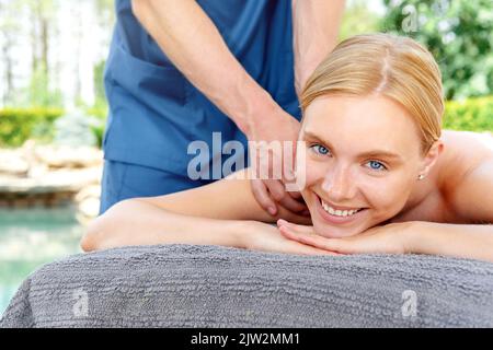 Bonne cliente blonde souriante et regardant la caméra pendant que le petit masseur se fait masser pendant la séance de spa sur le complexe Banque D'Images