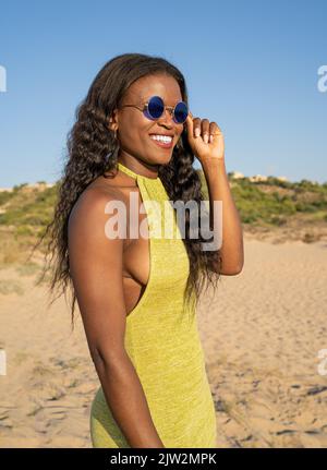 Bonne femme afro-américaine dans une robe élégante avec de longs cheveux ondulés prenant des lunettes de soleil et regardant loin avec le sourire tout en passant du temps sur la plage de sable Banque D'Images