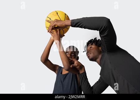 Le sportif afro-américain aide l'adolescent à apprendre le jeu du ballon pendant l'entraînement de basket-ball sur fond gris Banque D'Images