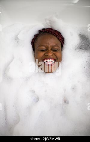 Vue de dessus d'une femme noire gaie riant avec les yeux fermés tout en étant allongé dans l'eau mousseuse dans la baignoire pendant la séance de spa à la maison Banque D'Images