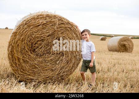 Un petit garçon mignon dans un t-shirt blanc et un short kaki est debout près des balles rondes de foin. Séance photo sur le terrain. Banque D'Images