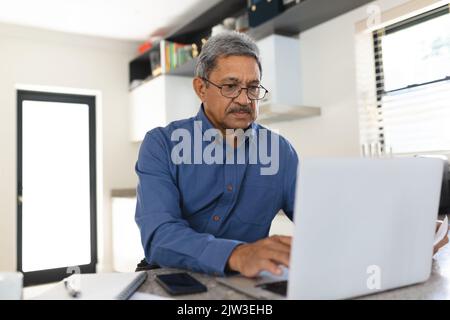 Homme de course mixte assis dans la cuisine à l'aide d'un ordinateur portable Banque D'Images