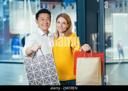 Jeune famille diverse asiatique homme et blonde femme shopping dans le supermarché, portrait de couple acheteurs regardant l'appareil photo et souriant et tenant des sacs colorés avec des marchandises et des achats. Banque D'Images