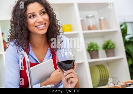 Belle heureuse hispanique Latina jeune femme ou fille avec des dents parfaites portant un tablier rouge buvant du vin rouge et utilisant une tablette d'ordinateur dans sa cuisine Banque D'Images