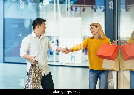 couple multiracial homme asiatique et femme caucasienne marchant ensemble dans le centre commercial après avoir fait du shopping avec des sacs de couleur entre les mains. passant devant les fenêtres du magasin avec des vêtements joyeux famille embrassant le sourire. Banque D'Images