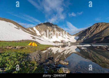 Vue sur les montagnes dans le nord du Canada pendant l'été avec lac calme et reflet de montagne dans l'eau en dessous avec tente jaune vif. Banque D'Images