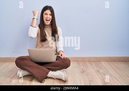 Jeune femme brune travaillant à l'aide d'un ordinateur portable assis sur le sol en colère et fou levant poing frustré et furieux tout en criant avec colère. r Banque D'Images