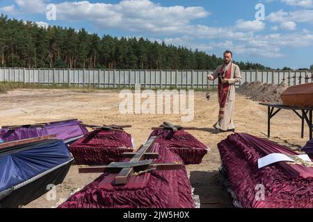 Un prêtre lit une prière et exécute une cérémonie funéraire pour les victimes innocentes de l'agression russe au cimetière de Bucha. Des rangées de cercueils avec des croix et le nombre de victimes non identifiées sont illustrés. Coffins contenant les corps de personnes non identifiées tuées dans le district de Bucha au moment de l'occupation russe, lors d'une cérémonie d'enterrement dans un cimetière de Bucha, région de Kiev, Ukraine. Des mois après le retrait des forces russes du district de Bucha, les corps qui n'ont pas été identifiés ont été enterrés et marqués de numéros dans un cimetière de Bucha. Banque D'Images