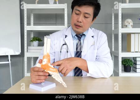Chirurgien orthopédique asiatique en robe blanche et stéthoscope pointant vers le modèle anatomique de l'articulation de l'os de la cheville du pied de squelette humain, présenté au patient par le biais d'une connexion sans fil Banque D'Images