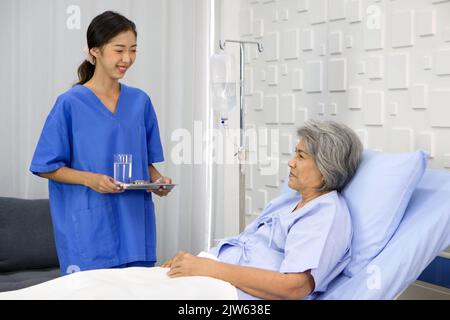 Une jeune infirmière asiatique en uniforme bleu apporte un médicament et un verre d'eau aux patients âgés à cheveux blancs. Une femme plus âgée étant traitée à l'hôpital, bien Banque D'Images
