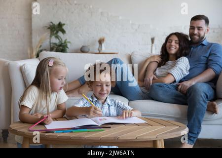 Les frères et sœurs d'avant-garde dessinant avec des crayons, les parents se reposant sur un canapé Banque D'Images