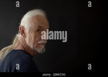 Portrait de studio de faible clé de beau vieux homme avec des cheveux gris dans une tresse regardant par-dessus son épaule à l'appareil photo. Horizontalement. Banque D'Images