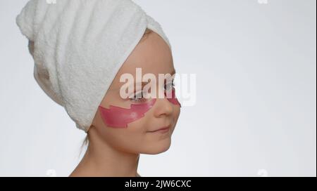 Belle jeune fille souriante dans une serviette sur la tête en appliquant des taches cosmétiques roses sous les yeux. Soin du visage pour enfant adolescent. Portrait de femme sur fond blanc. Un nettoyage naturel et parfait Banque D'Images