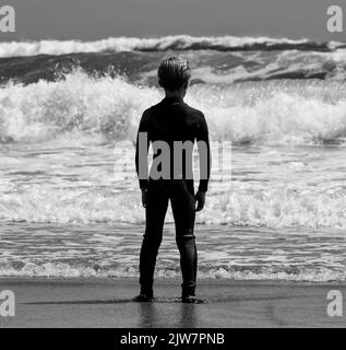 Jeune surfeur de lad en profitant des vagues à la plage de sable de Gwithian Cornwall. Noir et blanc. Prendre le temps de regarder la mer. Banque D'Images