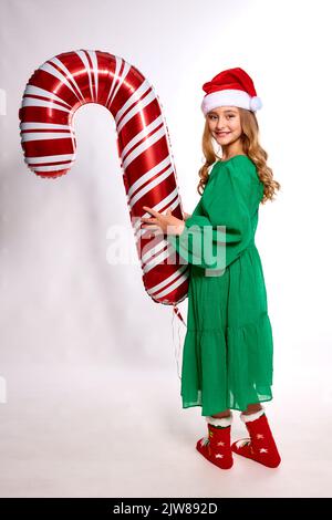 Fille de Noël en robe verte, chapeau de père noël et chaussettes chaudes rouges tient énorme ballon sous forme de canne dans ses mains Banque D'Images