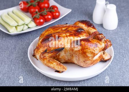 Poulet entier cuit à une délicieuse croûte. Un plat traditionnel. À l'arrière-plan se trouvent des concombres et des tomates cerises sur une assiette blanche. Gros plan. Banque D'Images