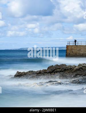 Vitesse d'obturation lente pour capturer les vagues douces qui se roulent jusqu'à la jetée de Porthleven, tandis qu'un pêcheur et des visiteurs explorent la jetée. Banque D'Images