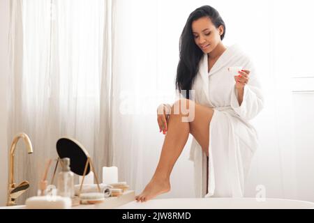 Jolie femme appliquant un hydratant sur les jambes debout dans la salle de bains moderne Banque D'Images