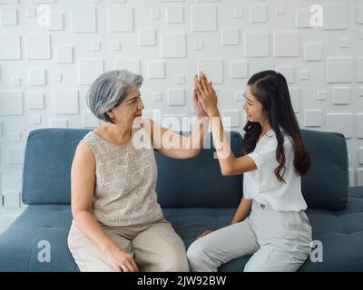 Femme asiatique âgée, maman donnant cinq hauts à sa fille gaie, jeune femme de beauté, assis sur un canapé gris dans la pièce blanche. Mère et fille Banque D'Images