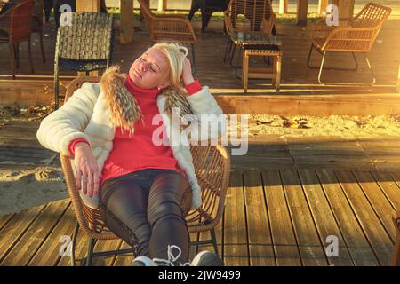Une femme dans un chandail et un manteau de fourrure est assise sur la terrasse dans une chaise en osier. Banque D'Images