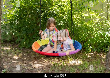 Deux enfants, filles, enfants assis sur une balançoire en plein air dans le jardin riant ensemble, s'amusant à l'extérieur. Activités extérieures, rires, vraies personnes Banque D'Images