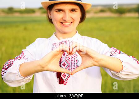 Recentrer le portrait de la jeune femme ukrainienne. Arrière-plan nature prairie. Montrer signe amour Ukraine. Fille ukrainienne souriante montrant l'amour gestuel. Vyshyvanka. Banque D'Images