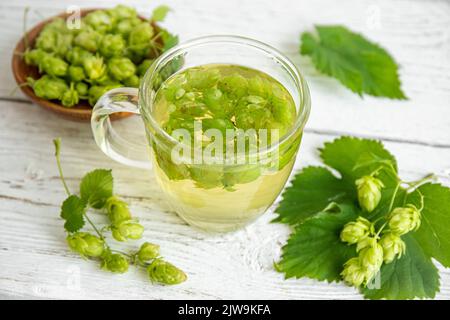 Boisson à base de thé à base de plantes à base d'Humulus lupulus, le houblon commun ou le houblon. Houblon fleurs avec tasse de thé sur fond de bois blanc, à l'intérieur de la maison. Banque D'Images