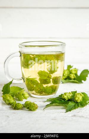 Boisson à base de thé à base de plantes à base d'Humulus lupulus, le houblon commun ou le houblon. Houblon fleurs avec tasse de thé sur fond de bois blanc, à l'intérieur de la maison. Banque D'Images