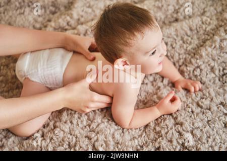 Femme mère effectuant un massage du dos à un bébé heureux allongé sur un couvre-lit beige. Maman masseuse et jeune garçon souriant. Enfant âgé d'un an et deux mois Banque D'Images