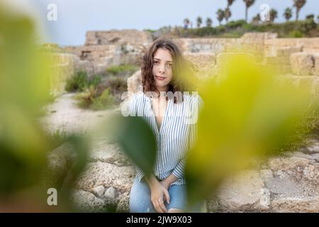 Une jeune fille de brunette pensive aux cheveux longs et bouclés dans une chemise en lin blanc avec des rayures bleues, un Jean bleu et des baskets vertes est posée sur la pierre dans le cadre Banque D'Images