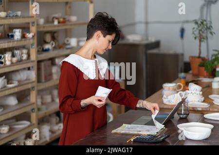 Young female entrepreneur pottery studio owner doing bookkeeping, paying income taxes Banque D'Images