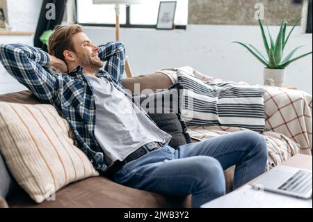 Détente à la maison. Satisfait positif calme élégant jeune homme caucasien, se reposant à la maison après une journée de travail, assis sur un canapé confortable, jetant ses mains derrière sa tête, regardant loin, souriant Banque D'Images