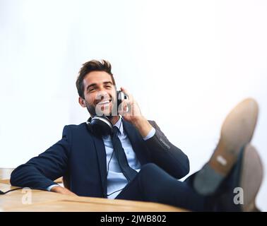 Les avantages d'être le patron. Un beau jeune homme d'affaires utilisant un téléphone portable avec ses pieds sur le bureau. Banque D'Images