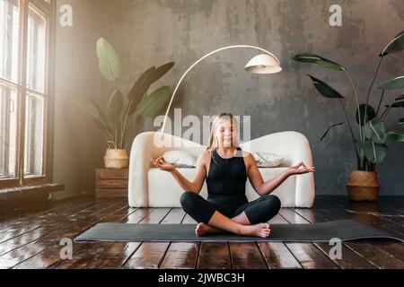 Jeune fille attrayante pratiquant le yoga, assis dans la moitié de l'exercice de Lotus, Ardha Padmasana pose, exercice, session de méditation, intérieur de maison. Quali élevé Banque D'Images