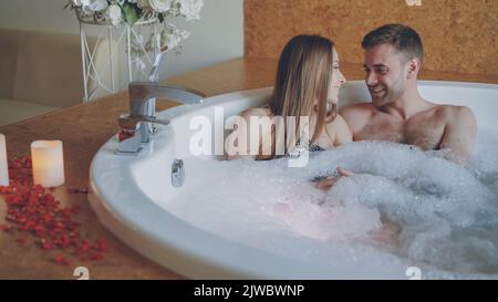 Les jeunes gens séduisants s'embrasent et parlent dans la baignoire bouillonnante chaude avec de la mousse dans le spa moderne. Relation romantique, conversation et concept de bien-être. Banque D'Images