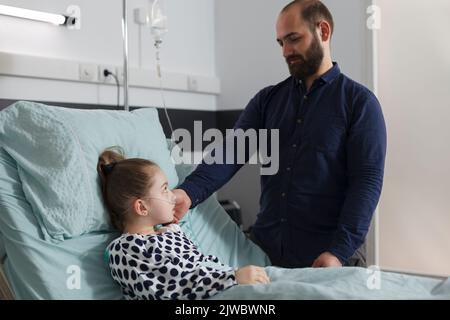 Parent attentionné prenant soin de la petite fille malade tout en touchant son visage. Un père aimant réconfortant une fille malade hospitalisée qui se repose dans le lit du patient dans la chambre de service pédiatrique de l'hôpital Banque D'Images