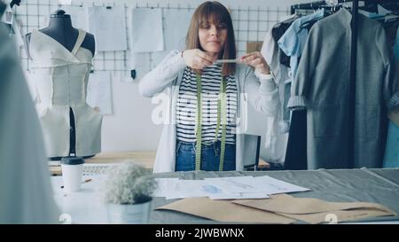 Belle jeune femme designer de mode prend des photos de croquis couchés sur la table. Elle fait des lays plats créatifs. Son studio est léger et plein d'articles de couture, d'outils et d'équipement. Banque D'Images