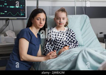 Portrait de la mère soignante réconfortant la fille malade hospitalisée se reposant dans le lit du patient. Femme assise à côté d'une petite fille malade dans la chambre d'un établissement de santé pour enfants tout en regardant la caméra Banque D'Images