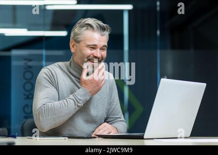 Homme d'affaires mûr et expérimenté, aux cheveux gris, travaillant dans un bureau moderne, regardant des vidéos amusantes et amusantes sur un ordinateur portable, le propriétaire de l'entreprise souriant et heureux dans les pauses de travail. Banque D'Images