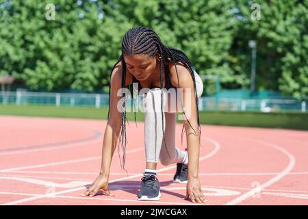 Une femme afro-américaine aux longues tresses prépare une course à pied à bas régime sur la piste rouge du stade Banque D'Images