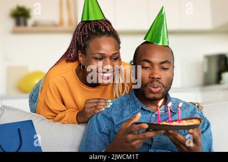 Homme afro-américain célébrant son anniversaire, en tenant une tarte savoureuse et en souhaitant avant de souffler les bougies allumées Banque D'Images