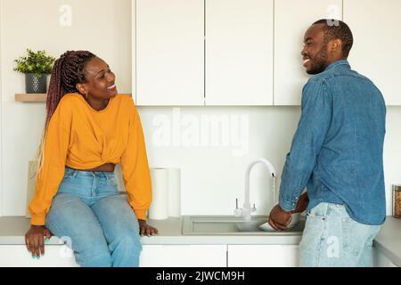 Jeune homme afro-américain lavant des plats et parlant à sa femme à l'intérieur de la cuisine Banque D'Images