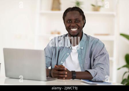 Jeune homme noir assis au bureau avec ordinateur portable et souriant à la caméra Banque D'Images