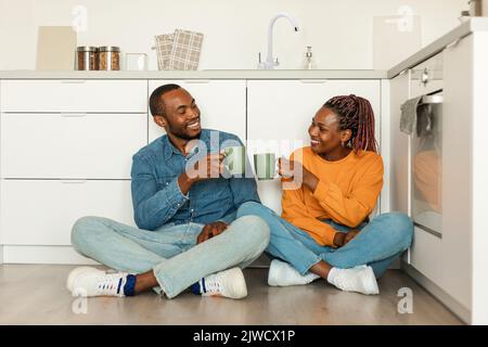 Romantique couple noir buvant du thé, assis dans la cuisine sur le sol et parlant, appréciant le matin à la maison Banque D'Images