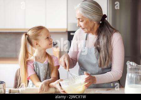 Une heureuse grand-mère européenne âgée en tablier enseignant à une petite fille de faire de la pâte, s'amuser ensemble dans la cuisine Banque D'Images