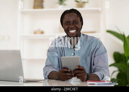 Happy Black Guy utilisant la tablette numérique tout en étant assis au bureau à la maison Banque D'Images