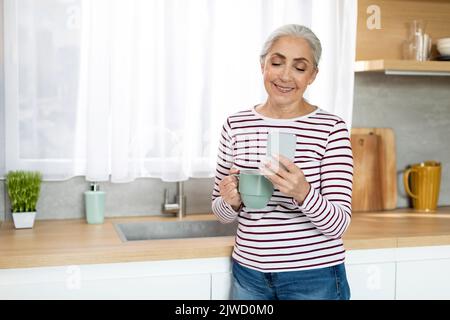 Communication mobile. Femme de chambre souriante se détendant dans la cuisine avec un téléphone intelligent et du café Banque D'Images